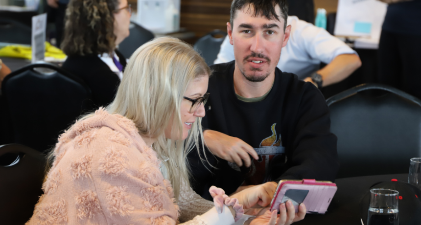 A woman wearing a pink jumper describing something on a mobile phone to a man with a disability