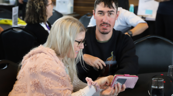 A woman wearing a pink jumper describing something on a mobile phone to a man with a disability