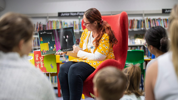 A library officer is reading a book to a group of children and their parrents